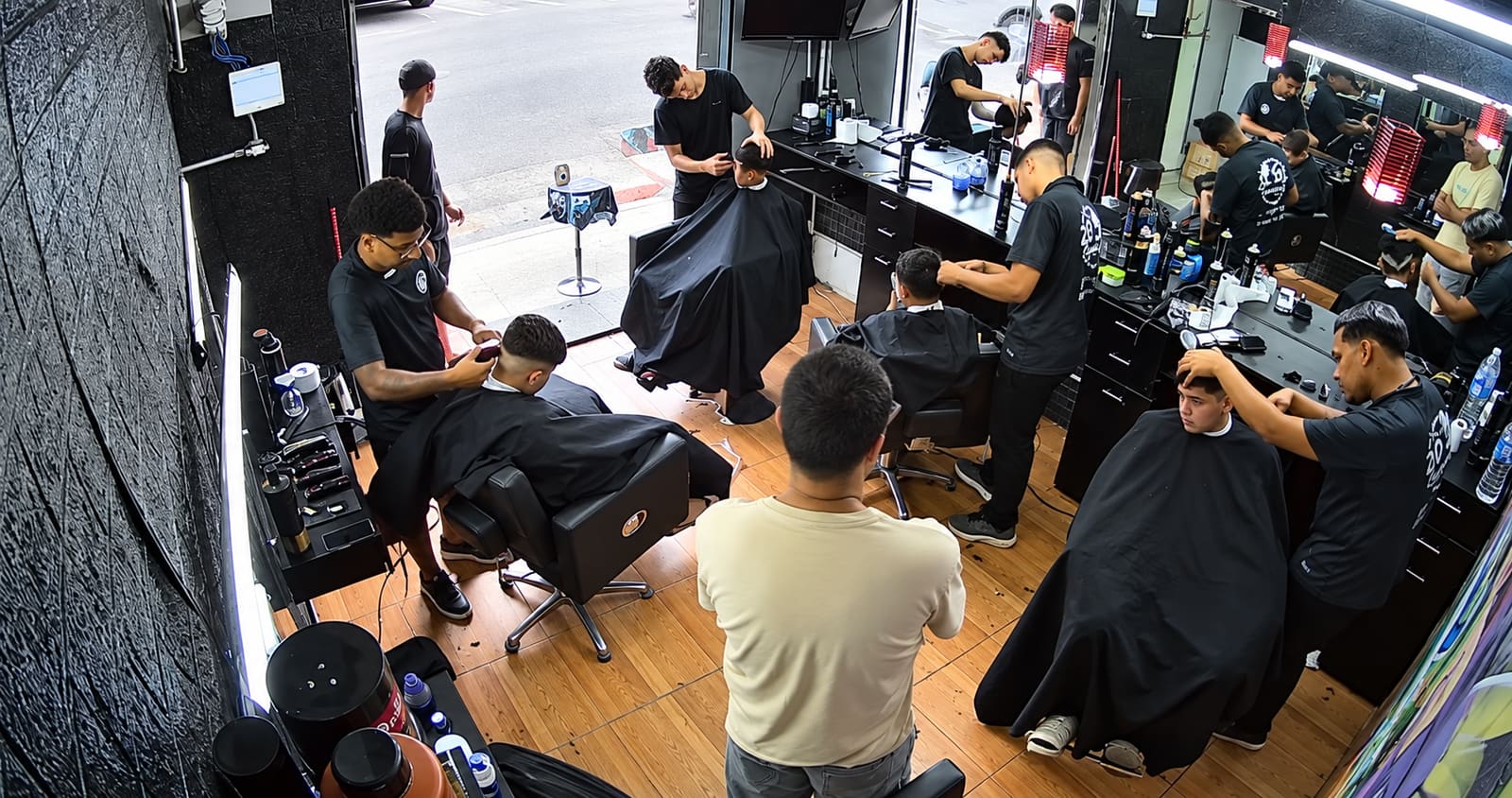 A 2D de casa cheia: barbeiros de uniforme preto atendendo clientes nas cadeiras, com a porta aberta para a rua